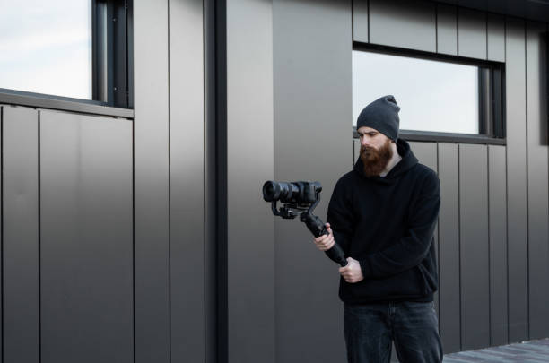 istock Bearded Professional videographer in black hoodie holding professional camera on 3-axis gimbal stabilizer. Filmmaker making a great video with a professional cinema camera. Cinematographer. 1251299484