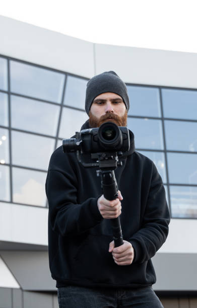 istock Bearded Professional videographer in black hoodie holding professional camera on 3-axis gimbal stabilizer. Filmmaker making a great video with a professional cinema camera. Cinematographer. 1251299528