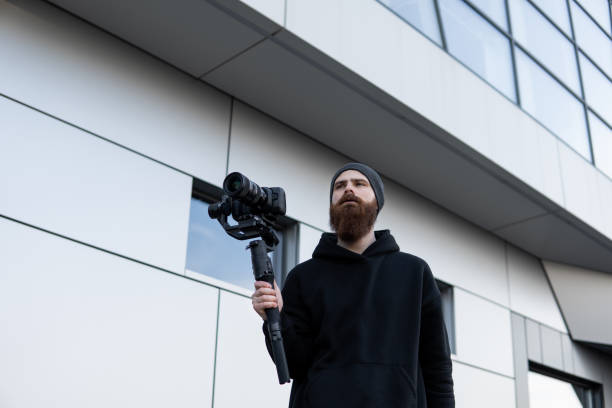 Bearded Professional videographer in black hoodie holding professional camera on 3-axis gimbal stabilizer. Filmmaker making a great video with a professional cinema camera. Cinematographer.