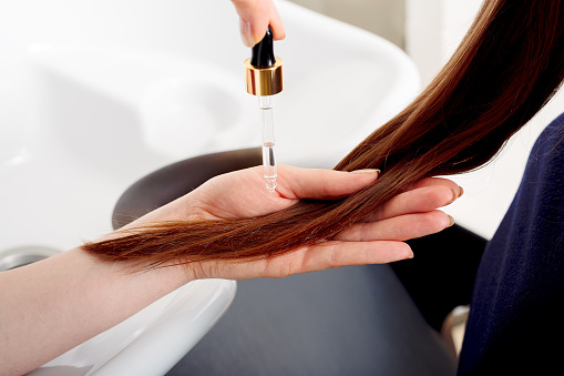 Female hands applying oil on long womans brown hair. Hair care cosmetics, bath beauty spa products. Female hair serum for treatment hair tips against beauty salon interior, bathroom.