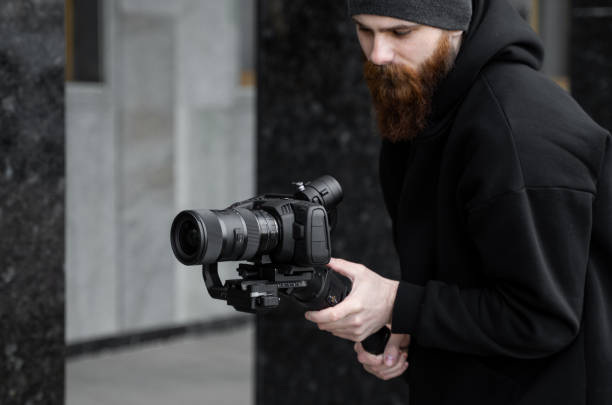 istock Bearded Professional videographer in black hoodie holding professional camera on 3-axis gimbal stabilizer. Filmmaker making a great video with a professional cinema camera. Cinematographer. 1251300918