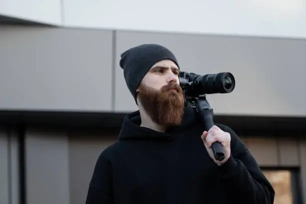 Bearded Professional videographer in black hoodie holding professional camera on 3-axis gimbal stabilizer. Filmmaker making a great video with a professional cinema camera. Cinematographer
