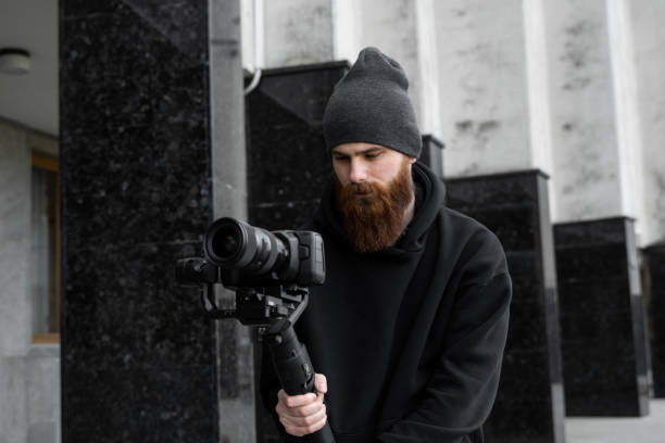 istock Bearded Professional videographer in black hoodie holding professional camera on 3-axis gimbal stabilizer. Filmmaker making a great video with a professional cinema camera. Cinematographer. 1251302071