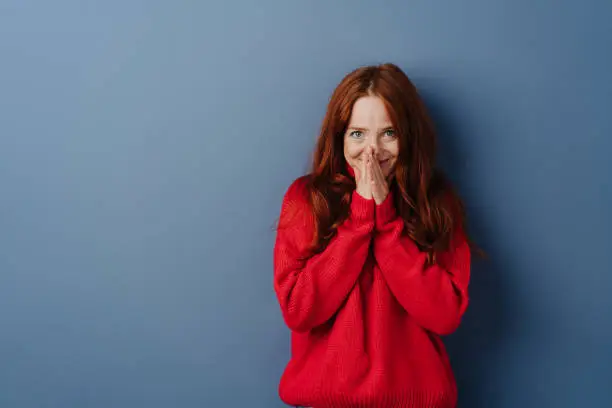 Cute young redhead woman with a shy smile holding her hands to her mouth as she looks at the camera over a blue studio background with copy space