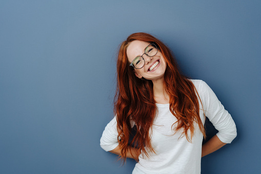 Happy attractive young redhead woman with sweet vivacious smile and tilted head over a blue studio background with copy space