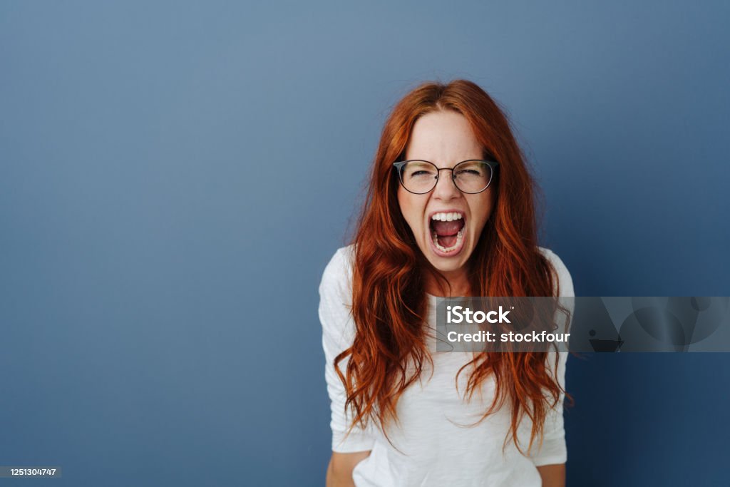 Angry young woman throwing a temper tantrum Angry young woman throwing a temper tantrum yelling at the camera with a furious expression over a blue studio background with copy space Shouting Stock Photo