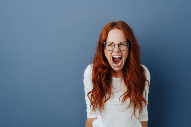 Angry young woman throwing a temper tantrum Angry young woman throwing a temper tantrum yelling at the camera with a furious expression over a blue studio background with copy space shouting stock pictures, royalty-free photos & images