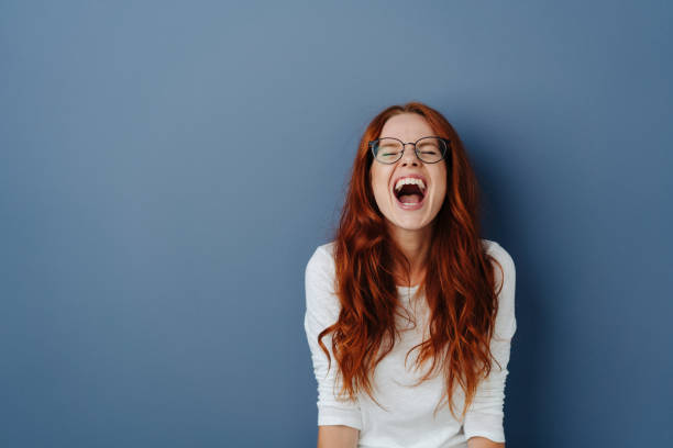 Young woman enjoying a loud guffaw Young woman with long red hair and glasses enjoying a loud guffaw or belly laugh over a blue studio background with copy space mouth open human face shouting screaming stock pictures, royalty-free photos & images