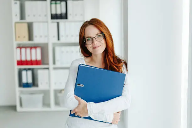 Young businesswoman standing looking at camera with a quiet confident friendly smile while clutching a large office binder
