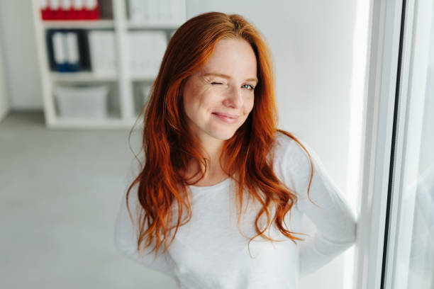 Cute playful young woman winking at the camera Cute playful young woman winking at the camera with a mischievous grin as she waits at a door to the office winking stock pictures, royalty-free photos & images