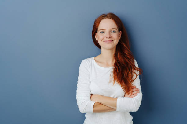 Pleased young redhead woman with a beaming smile Pleased confident young redhead woman with a beaming smile and folded arms posing on a blue studio background with copy space grinning at the camera 30 34 years stock pictures, royalty-free photos & images