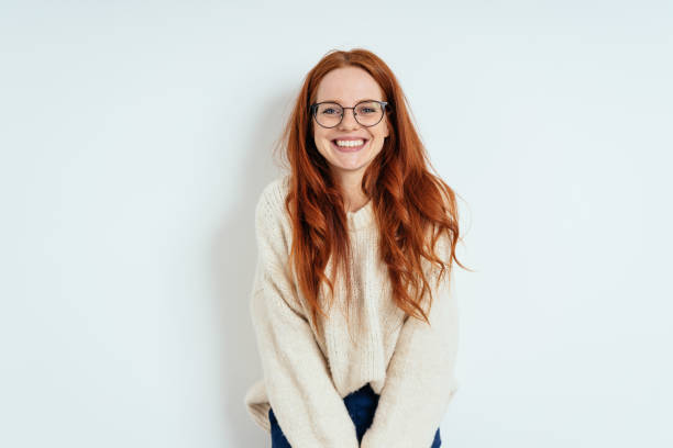 Smiling friendly young woman wearing spectacles Smiling friendly young woman with long red hair wearing spectacles looking at the camera with a vivacious smile against a white interior wall with copy space red hair stock pictures, royalty-free photos & images