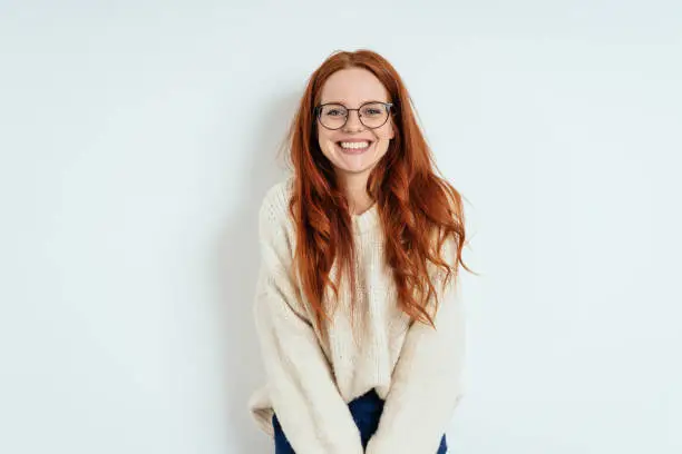 Smiling friendly young woman with long red hair wearing spectacles looking at the camera with a vivacious smile against a white interior wall with copy space