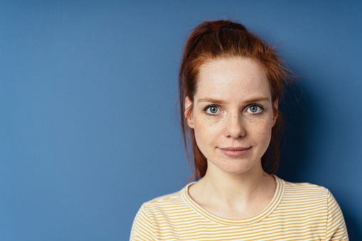 Pretty young redhead woman with large blue eyes smiling quietly at the camera in a close up portrait on a blue studio background with copy space