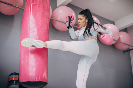 One fit young woman with boxing gloves kicking a punching bag in the gym.
