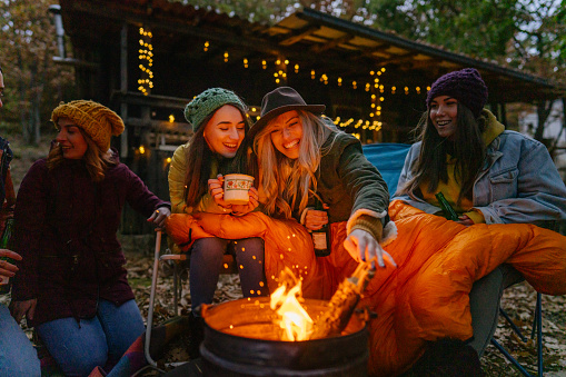 Photo of a couple of smiling friends sitting by the campfire on a cool autumn evening; enjoying each other's company.