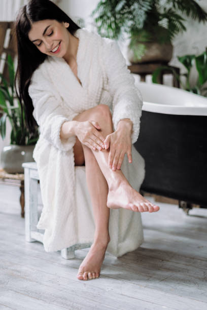 Young adult woman spending free time at home Happy young adult woman making massage, apply cream on skin, using organic cosmetic, skincare and bodycare product. Girl in bathrobe spending morning at bathroom, sitting on chair near bath epilator stock pictures, royalty-free photos & images