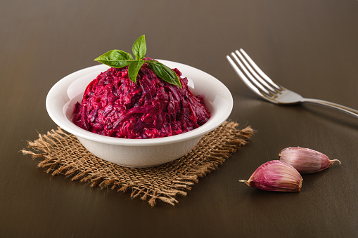 Grated boiled beet salad with garlic and mayonnaise in a white bowl on a brown table. Side dish and vegetarian garnish. Russian cuisine. Front view.