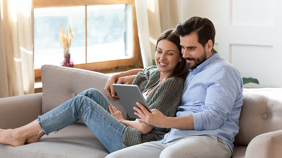 Happy young couple relaxing on cozy couch, using computer tablet together, smiling beautiful woman and man hugging, looking at device screen, watching movie or video, shopping online, browsing apps