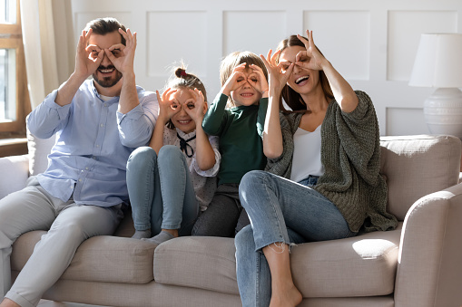 Family portrait happy parents with children making funny faces, posing on cozy couch at home, mother and father with kids binoculars glasses eyewear shape gesture, looking at camera through fingers