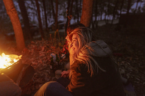 Photo of two female friends sitting by the campfire on a cool autumn evening in the woods; keeping warm during the cool night.