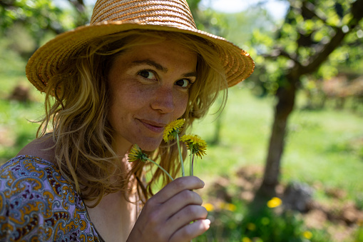 A pretty blonde woman in a paisley dress and a hat is smiling while smelling dandelions in a garden in spring