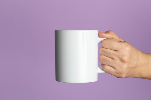 Caucasian female is holding a white coffee mug in hand in front of a purple background.