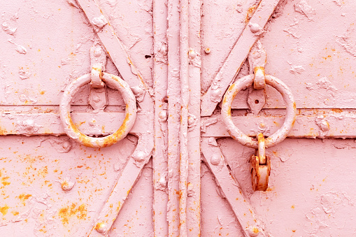 Fragment of a closed old metal door or gate with round knockers and a rusty padlock. Closeup shabby worn texture of iron with pieces of peeling pink paint layer with spots of red rust. Background.