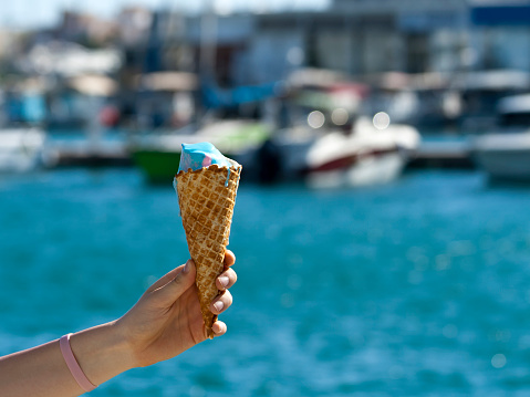 Ice cream waffle cone in hand in front of blurred blue sea and boats