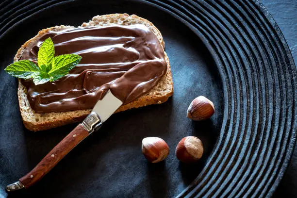 Chocolate cream spread on bread slice and hazelnuts on a dark black rustic plate with mit leaves on top