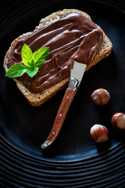Chocolate cream spread on bread slice and hazelnuts on a dark black rustic plate with mit leaves on top