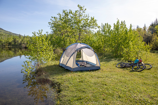 Tent and two e-bikes stand on sunny meadow by lake and Rocky Mountains