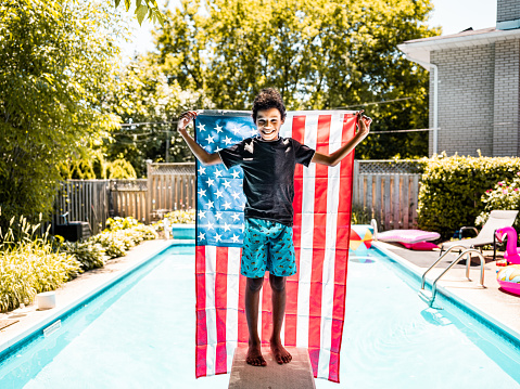 Black boy holding American National flag while standing on a diving board by the backyard pool.