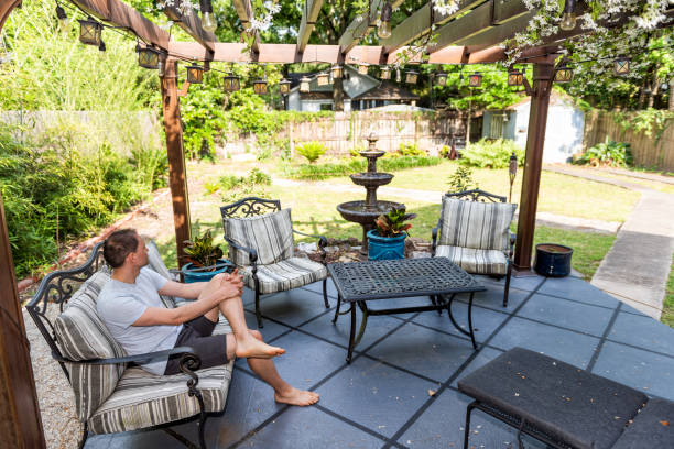 Young man sitting on patio lounge chair in outdoor spring flower garden in backyard of home zen with water fountain, pergola canopy gazebo, table, plants, sofa Young man sitting on patio lounge chair in outdoor spring flower garden in backyard of home zen with water fountain, pergola canopy gazebo, table, plants, sofa gazebo photos stock pictures, royalty-free photos & images