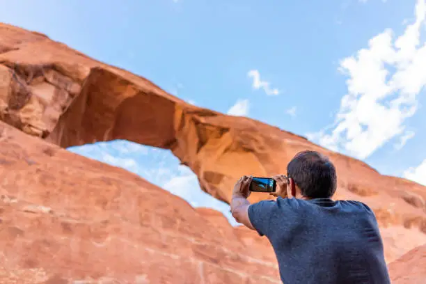 Photo of Arches National Park canyons with Skyline Arch in background and young man standing taking picture of view with phone on trail hike in Utah, USA