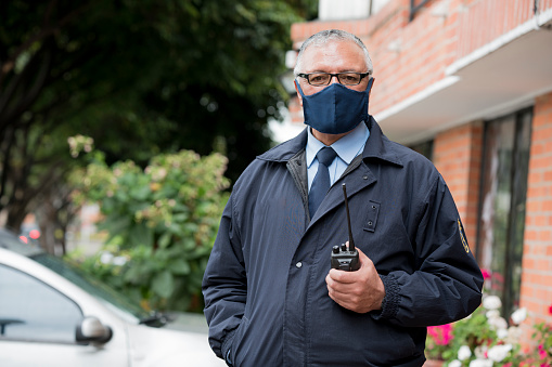 The doorman of the building is a Latino from Bogota Colombia, between 50 and 59 years of age, looking at the camera in a portrait while wearing his mask to avoid the Covid 19 and holding his radio