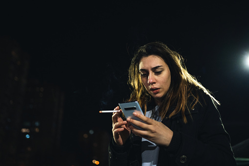 Woman standing in a parking lot, smoking a cigarette while using her phone