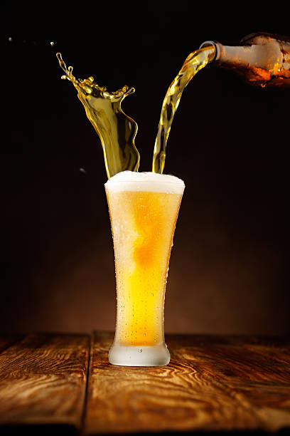 Splashing - Beer pour in glass Pouring beer. Splashing. Ice Cold Bottle and Glass of  Beer , covered with water drops - condensation.Glass standing on wooden table with dark gradient background. Foam on the beer looks real and perfect while the colors between the glass and the bottle fit perfectly with the contrast and shapes on the table. beer glass splash stock pictures, royalty-free photos & images