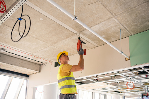 A man using an electric pneumatic drill makes a hole in the wall, a professional construction worker with a safety hard hat, gloves. inside the construction site