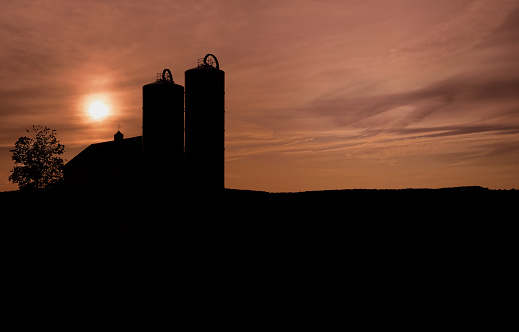 Two grain farm silos with a dramatic orange sky and the sun going down just over the buildings.