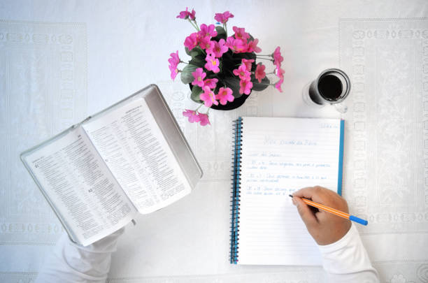 Hands of a mid age Brazilian woman in a living room, holding a Bible in Brazilian Portuguese and a pen, reading it and making study notes of the book of Psalms on her notebook. On the table there are a coffee cup and a vase with pink flowers in it.