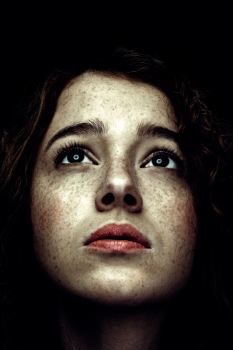 A dark, high contrast image of a mysterious beautiful young woman, looking up into the distance with an expression of intrigue, desire, hope, or longing.   Vertical with copy space.