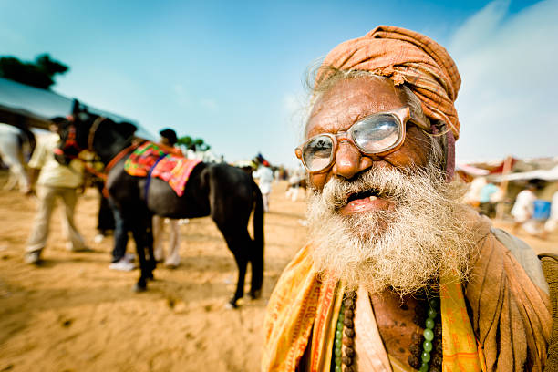 Eccentric Indian Man Pushkar Camel Fair India Character Portrait Eccentric Indian Man with white beard and thick old glasses in traditional indian clothing with turban at Pushkar Fair Ground. Horses and Camel Traders of Pushkar Fair in the background. Pushkar, Rajasthan, India. india indian culture market clothing stock pictures, royalty-free photos & images