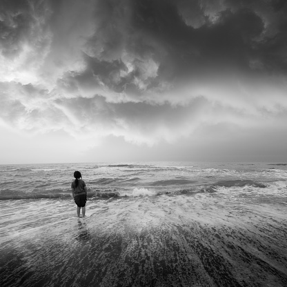 A child on the beach, standing in a foaming sea wave, looking at aproaching storm. Black and white photo. 