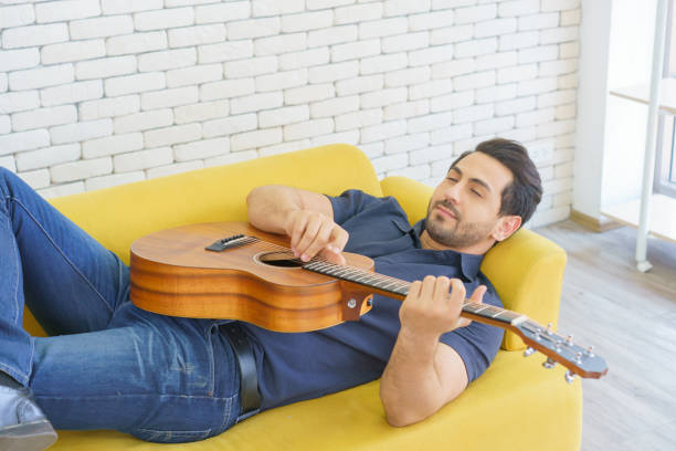 happy man playing guitar while sitting on sofa in living room, enjoying carefree time at home. - 16710 imagens e fotografias de stock