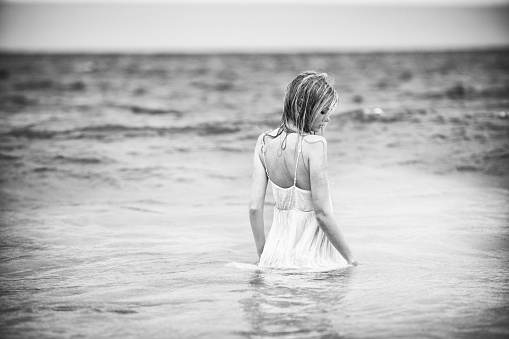 Beautiful blond young woman walking out into ocean, looking back over her shoulder. Copy space, black and white.