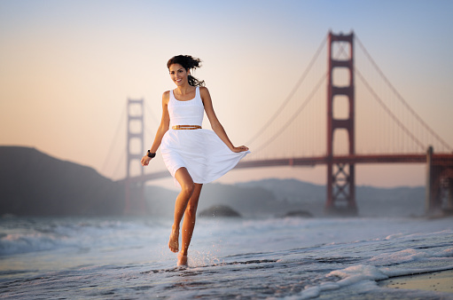 Beautiful model running through the surf on Baker Beach in  front of the Golden Gate Bridge at sunset. Warm sun coming from the left. Dusk building up in front of the Golden Gate Bridge in the background. Great natural playful Expression!