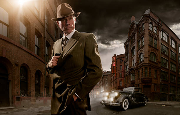 1940's black gangster 1940's stylised image of man in suit and fedora hat standing in run down city with vintage car in background about to take out gun from pocket film noir style photos stock pictures, royalty-free photos & images