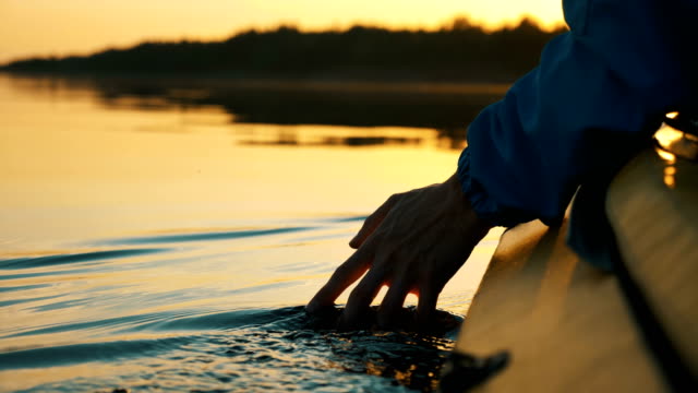 unity with nature, man puts his hand into the water while sitting in kayak against golden sunset, camera slow motion