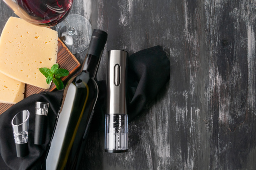 Gray electric corkscrew made of metal. Lies next to a bottle of wine, aerators and a vacuum stopper on black fabric. Next to a glass of wine and a piece of cheese. Dark wooden background. View from above.